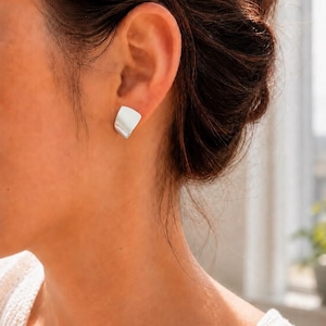 May include: A small, silver-toned stud earring with a smooth, curved, fan-like shape. The earring features a polished, reflective metallic surface that catches the light, creating a subtle sheen across its slightly flared, rounded edges.