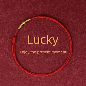 May include: Red string bracelet with a gold accent and the word "Lucky" in the center. The text "Enjoy the present moment" is below the word "Lucky". The bracelet is on a red background.