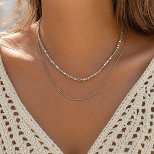 May include: A layered necklace set consisting of two silver-toned chains. The shorter chain features a series of small, uniform square-shaped beads, while the longer chain is a simple, smooth, and thin snake chain that rests below the beaded strand.