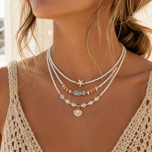 May include: A multi-strand necklace set with marine-themed pendants. The top strand features a starfish, the middle a small fish, and the bottom a seashell pendant with a pearl. The necklaces are white and gold in colour.