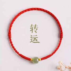 May include: A red braided bracelet featuring a green stone bead and gold details. The background displays the Chinese characters for "Turn Luck".