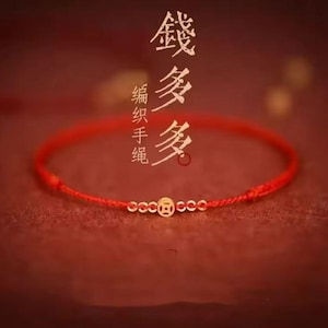 May include: A red string bracelet with gold-coloured beads and a central coin-shaped charm. The bracelet is set against a blurred red background with Chinese characters.