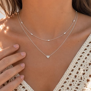 May include: A double-layered silver necklace featuring a delicate chain with small cylindrical beads on the upper strand and a simple, smooth silver heart charm centred on the lower strand, creating a layered look around the neck.