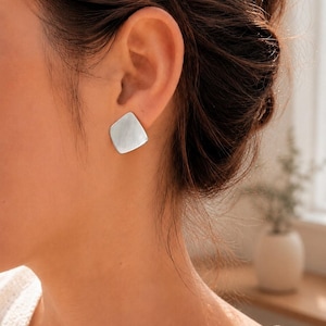 May include: A silver-toned stud earring with a smooth, square-shaped design featuring rounded corners and a flat, reflective surface. The earring is positioned securely on the earlobe, showcasing a clean, metallic finish and a simple geometric form.