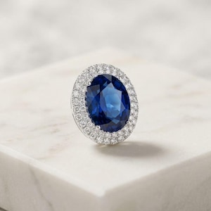 May include: A striking ring featuring a large, oval-cut, deep blue gemstone, surrounded by a halo of smaller, clear stones. The ring is set in a silver or white metal, resting on a white marble surface. The gemstone's colour and cut are the main features.