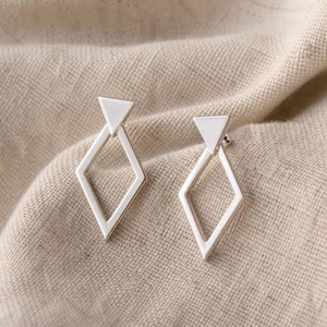 May include: A pair of silver geometric earrings. Each earring has a triangular stud above a diamond-shaped frame. The earrings are presented on a neutral fabric background, highlighting their minimalist design and modern aesthetic.