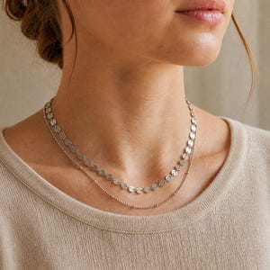 May include: A silver necklace with two strands. The top strand has small, flat, circular pendants. The bottom strand is a delicate chain with tiny beads. The jewelry is worn on a person wearing a beige jumper.