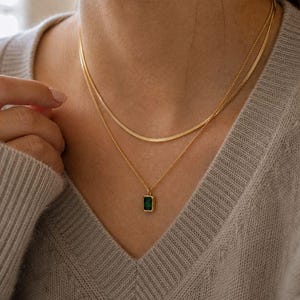 May include: Two gold necklaces. The top necklace is a thin, flat chain. The bottom necklace has a rectangular, emerald-green pendant. The necklaces are worn on a person wearing a grey jumper.