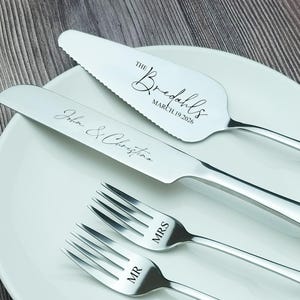 Puede incluir: Un juego de servir pastel de boda de acero inoxidable y tenedores se muestran en un plato blanco. El juego incluye un servidor de pastel, un cuchillo para pastel y dos tenedores. El servidor y el cuchillo están grabados con nombres y una fecha. Los tenedores están grabados con "MR" y "MRS".