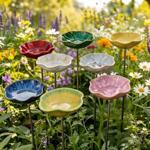 Puede incluir: Un conjunto de ocho bebederos para pájaros de cerámica, cada uno con forma de flor de bordes ondulados y montado sobre una fina estaca de metal. Los cuencos presentan varios acabados brillantes, incluyendo rojo, verde oscuro, amarillo, azul, rosa y blanco moteado.