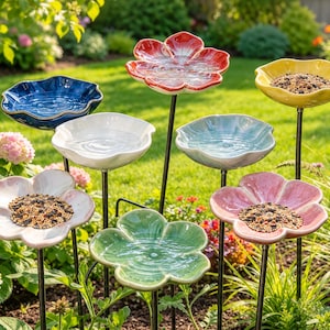 May include: A collection of eight ceramic flower-shaped bird feeders and baths on thin black metal stakes. The dishes feature scalloped edges in red, yellow, blue, white, light blue, pink, and green, with some containing birdseed mixes.