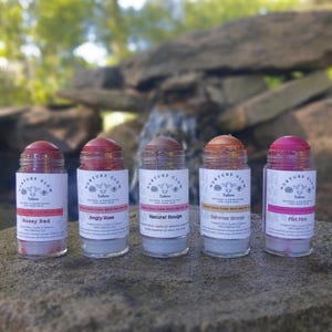May include: Five Tallow Glow color sticks in a row, each with a different shade of color. The colors are Rosey Red, Angry Rose, Natural Rouge, Summer Bronze, and Flirt Pink. Each stick has a white label with the brand name and product information.