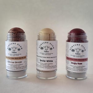 May include: Three Tallow Glow Color Sticks in clear glass jars. The sticks are labeled "Summer Bronze", "Snow White", and "Angry Rose". Each jar has a white label with a cow illustration and the words "Nurture Glow". The sticks are 3 oz.