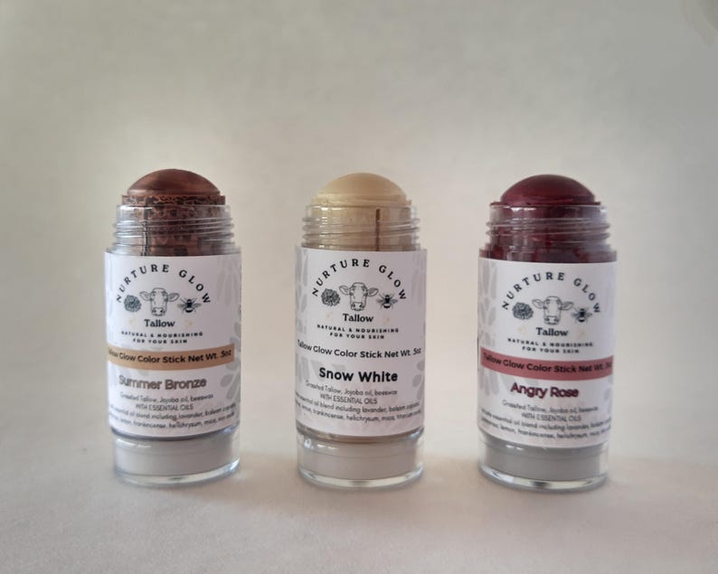 May include: Three Tallow Glow Color Sticks in clear glass jars. The sticks are labeled "Summer Bronze", "Snow White", and "Angry Rose". Each jar has a white label with a cow illustration and the words "Nurture Glow". The sticks are 3 oz.