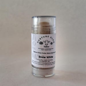 以下が含まれることがあります： 「Nurture Glow」の「Snow White」のタローグローカラースティックの透明なチューブ。ラベルには牛のイラストとテキストが描かれています。製品には、タロー、ホホバオイル、ミツロウ、エッセンシャルオイルが含まれています。正味重量30g。