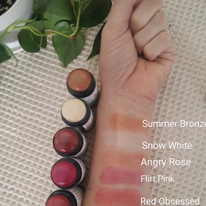 May include: A collection of five makeup sticks in various shades, including Summer Bronze, Snow White, Angry Rose, Flirt Pink, and Red Obsessed. The colors are swatched on a wrist, with the sticks lined up beside them. The sticks have black bases and white labels.