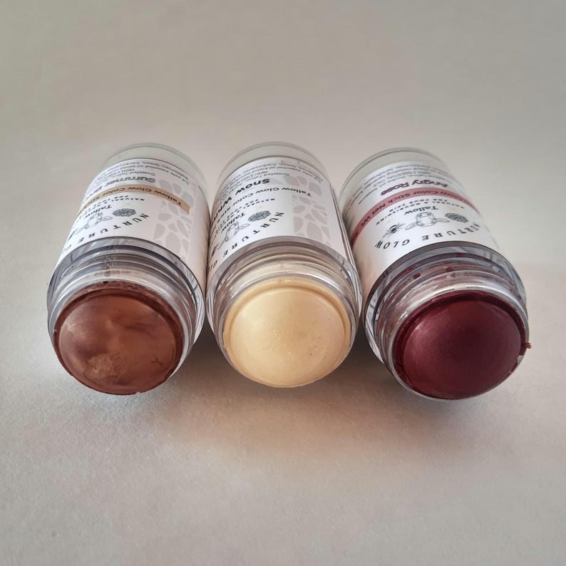 May include: Three clear tubes of tallow glow color sticks. The sticks are in shades of brown, cream, and red. Each tube has a white label with black text and a floral design. The tubes are arranged on a white surface.