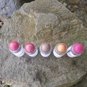 May include: Five small, clear jars with colorful lip balm. The balm shades range from pink to bronze. The jars are arranged on a large, gray rock, with a stream in the background. Each jar has a white label.
