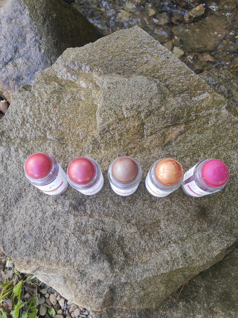 May include: Five small, clear jars with colorful lip balm. The balm shades range from pink to bronze. The jars are arranged on a large, gray rock, with a stream in the background. Each jar has a white label.
