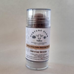 以下が含まれることがあります： サマーブロンズカラーのスティックが入った透明なプラスチックチューブ。ラベルには、牛と蜂のイラストとともに「Nurture Glow Tallow」と書かれています。テキストには、エッセンシャルオイルと製品の正味重量も記載されています。