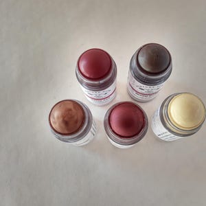 May include: Five cosmetic sticks with various shades of color. The sticks are in clear glass containers with white labels and red, brown, and yellow tops. The sticks are arranged on a white surface.