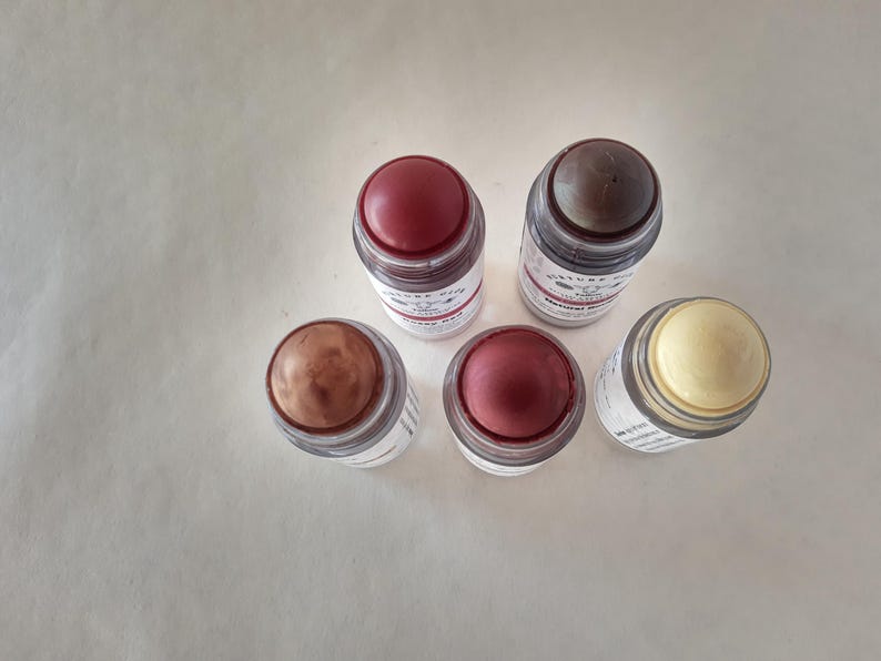 May include: Five cosmetic sticks with various shades of color. The sticks are in clear glass containers with white labels and red, brown, and yellow tops. The sticks are arranged on a white surface.