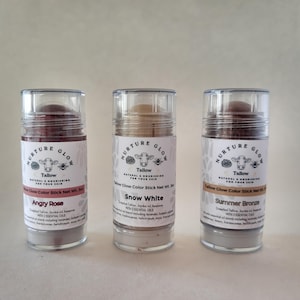 May include: Three cylindrical cosmetic sticks labeled "Nurture Glow Tallow" in a row. The sticks are "Angry Rose", "Snow White", and "Summer Bronze". Each stick has a clear cap and a label with a cow illustration and text.