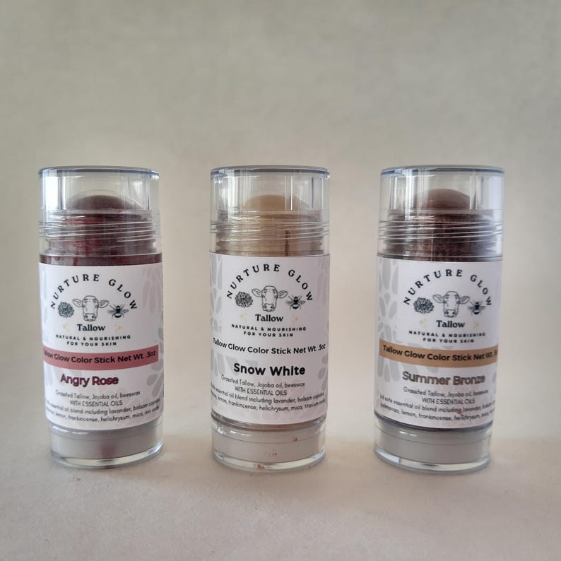 May include: Three cylindrical cosmetic sticks labeled "Nurture Glow Tallow" in a row. The sticks are "Angry Rose", "Snow White", and "Summer Bronze". Each stick has a clear cap and a label with a cow illustration and text.