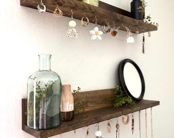 Könnte beinhalten: Zwei Holzregale mit Haken zum Aufhängen von Schmuck. Das obere Regal hat ein gerahmtes Bild, ein Teeglas und eine Vase mit Grünpflanzen. Das untere Regal hat eine Glasflasche mit Grünpflanzen, einen Holzdiffusor und einen Spiegel. An den Haken hängen Ohrringe und Halsketten.