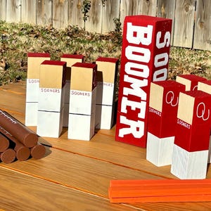 May include: A collection of wooden game pieces and decorative blocks. The blocks are painted red, white, and tan, with the word "BOOMER" and "SOONERS" displayed. Brown cylindrical pieces and an orange rectangular piece are also present.