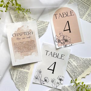 May include: Three wedding table cards with floral illustrations and the number 4. One card reads "TABLE 4" with a blush pink arch design. Another card says "CHAPTER I: How we met" with text. The cards are on a white surface.