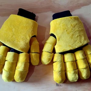 Op de afbeelding: Een paar felgele pluche handschoenen met zwarte manchetten. De handschoenen hebben afzonderlijke vingersecties, waardoor ze een cartoonachtig uiterlijk hebben. De handschoenen zijn gemaakt van een zacht, getextureerd materiaal.