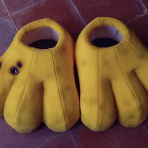 Op de afbeelding: Een paar felgele, oversized handvormige pantoffels. Elke pantoffel heeft een grote opening voor de voet en drie prominente vingerachtige verlengingen. De pantoffels hebben een donkere vlek aan de bovenkant. Op een roodbruin oppervlak.
