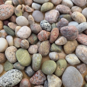 May include: A collection of smooth, rounded river stones in various sizes and colors, including shades of gray, tan, white, pink, and green. The stones have diverse textures, ranging from speckled and mottled patterns to solid, muted earth tones.