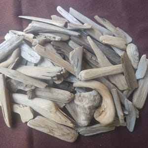 May include: A pile of weathered driftwood pieces in various shapes and sizes. The wood is primarily light gray and beige, with some pieces showing darker, weathered tones. The pieces are arranged on a dark brown surface.