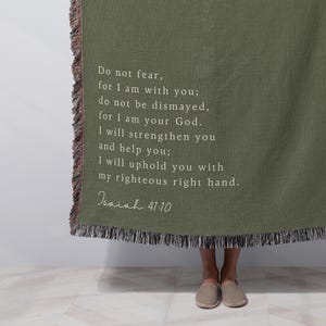 May include: Olive green throw blanket with fringed edges. The blanket features white text with a quote from Isaiah 41:10. The blanket is held up by a person.
