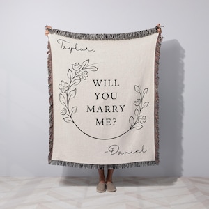 May include: Cream-colored woven blanket with black text and floral design. The text reads "Taylor, WILL YOU MARRY ME? -Daniel". The blanket has a fringed edge in shades of brown and black. A proposal-themed gift.