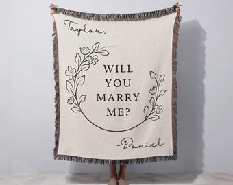 Pode incluir: Cobertor tecido creme com texto preto e design floral. O texto diz "Taylor, WILL YOU MARRY ME? -Daniel". O cobertor tem uma borda com franjas em tons de marrom e preto. Um presente com tema de proposta.