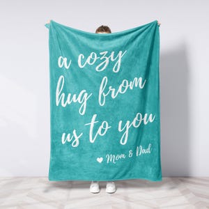 May include: A teal blanket with white script that reads "a cozy hug from us to you". Below the text is "Mom & Dad" with a heart symbol. The blanket is held up by a person wearing white sneakers.