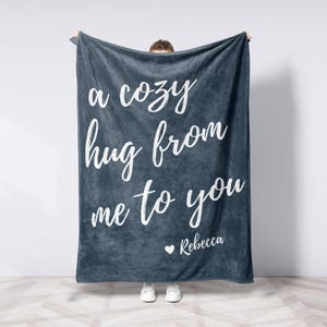 May include: A dark blue blanket with the text "a cozy hug from me to you" in white script. The name "Rebecca" is written below the text, with a small heart symbol. The blanket is held up by a person.