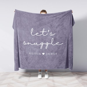 May include: A large, lavender blanket with the words "let's snuggle" in white script. Below the text are the names "Olivia" and "James" separated by a white heart symbol. The blanket is held up by a person wearing white trainers.