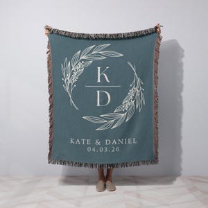 Könnte beinhalten: Türkisfarbene Decke mit weißem floralem Kranzdesign und Fransenbesatz. In der Mitte befinden sich die Initialen "K" und "D" über den Namen "KATE & DANIEL" und dem Datum "04.03.26".
