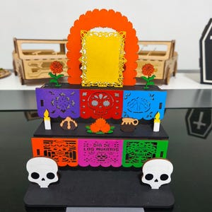 Puede incluir: Un colorido altar del Día de Muertos con un marco amarillo central, detalles naranjas y banderines decorativos recortados. El altar presenta calaveras, velas y elementos florales, con el texto "Día de los Muertos".