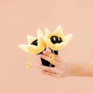 May include: Two handmade crochet flower-shaped keychains. The flowers are yellow with black centers and gold-colored key rings. The stems are black. The keychains are held against a peach-colored background.
