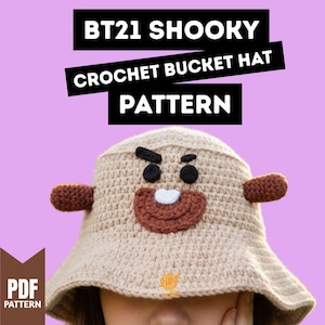 May include: A beige crochet bucket hat featuring the character Shooky from BT21. The hat has brown ear-like protrusions, a brown mouth, black eyes, and a white tooth. The text "BT21 SHOOKY CROCHET BUCKET HAT PATTERN" is displayed above the hat.