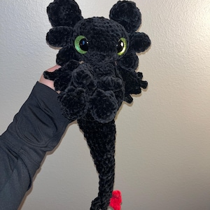 May include: A handmade black plush dragon with large green eyes and a red tail fin. The dragon is made of a soft, textured material and has large, rounded ears. A unique and cuddly plush toy.