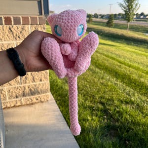 May include: A light pink plush toy crafted from soft, textured yarn. The creature features a rounded head with pointed ears, large bright blue eyes, small arms held near the chest, and a long, slender tail extending downward from the body.