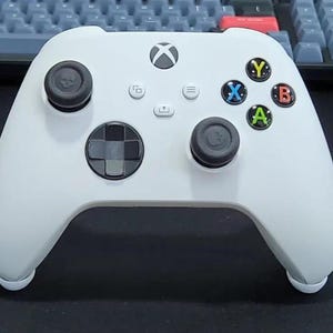 May include: A white Xbox controller with black thumbsticks and a directional pad. The controller features colored buttons (green, red, blue, yellow) and a central Xbox logo. The controller has a sleek, ergonomic design.
