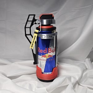 May include: A custom Red Bull can holder with a black handle and a red base. The can is blue and silver with the Red Bull logo and text. A yellow tag reads "GIVES YOU WINGS".