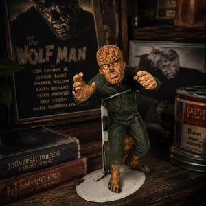 Peut inclure: Figurine du Loup-Garou, personnage de film d'horreur classique, sur une étagère en bois. La figurine présente une peau texturée et une tenue verte. Une affiche de film encadrée et des boîtes de film vintage évoquent le thème de l'horreur.